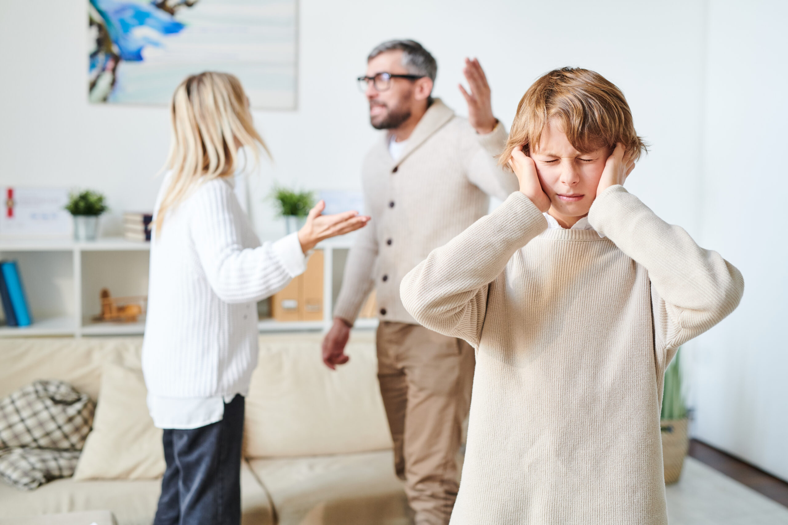 Distraught teenage son in sweater suffering from parents conflict: he keeping eyes closed and plugging in ears with fingers while his nervous parents fighting in background
