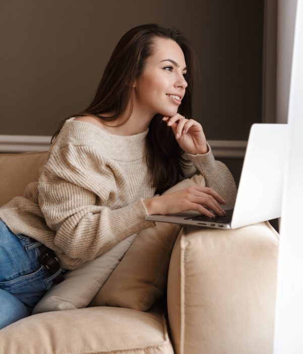 Image of joyful caucasian woman smiling and using laptop while sitting on couch in living room